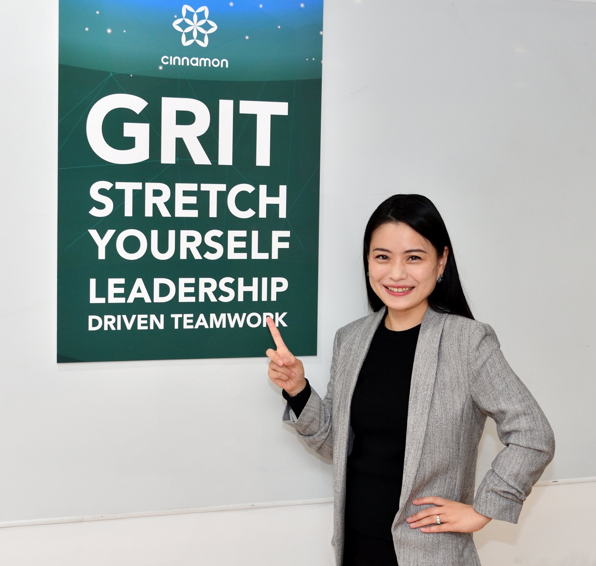 Miku Hirano, CEO of Cinnamon Inc, standing in front of a sign that reads Grit, Stretch Yourself, Leadership Driven Teamwork